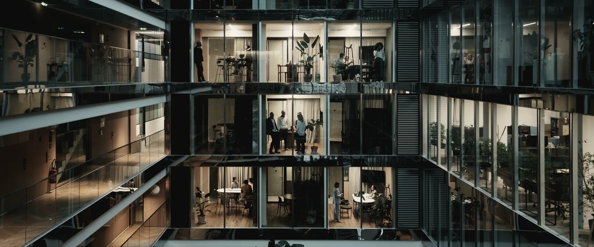 Teams at work in the lit offices of a glass atrium at dusk